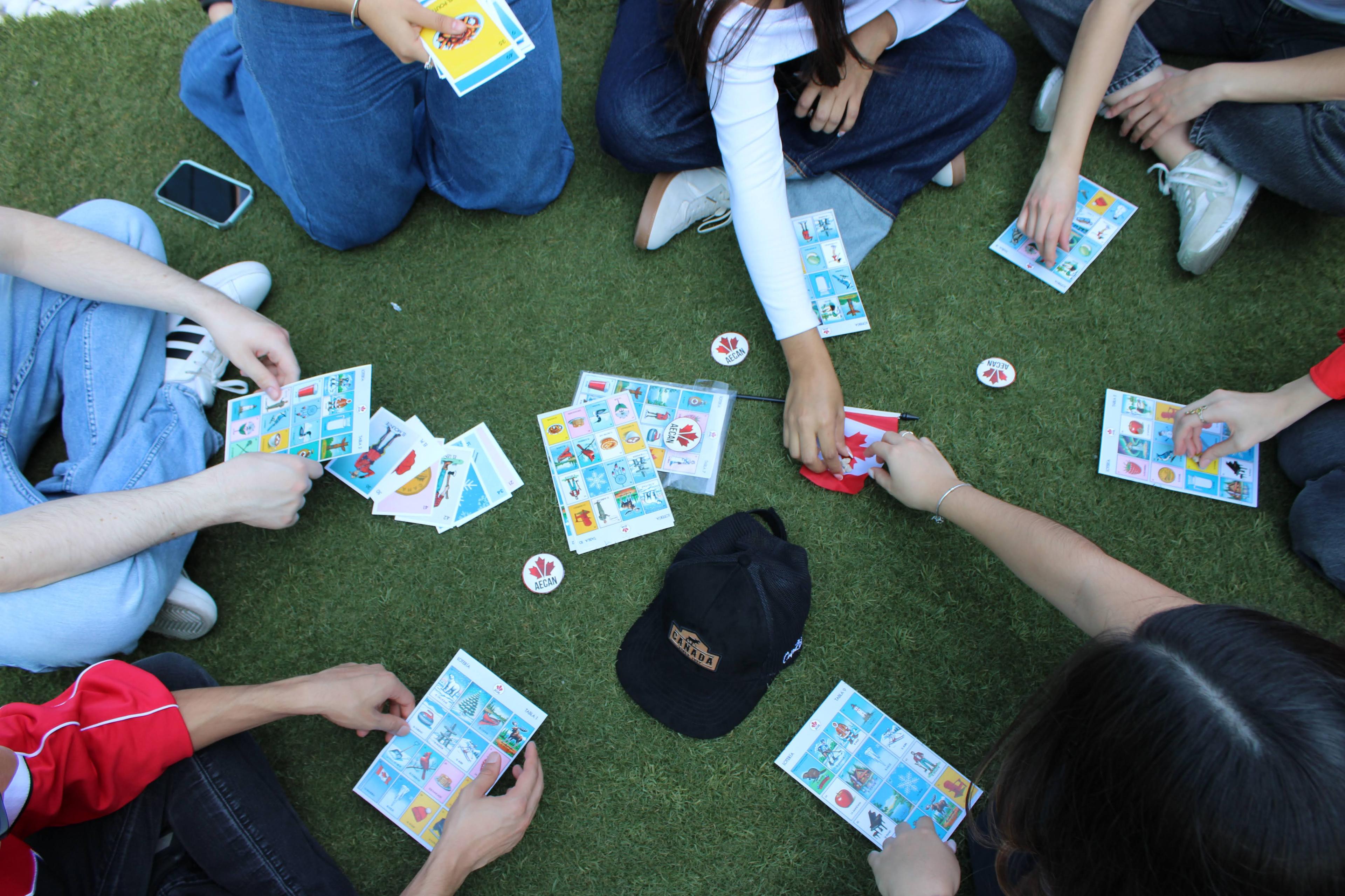 Top-down view of students sitting in a circle on campus turf playing Lotería Canadiense, tablas and AECAN tokens spread out