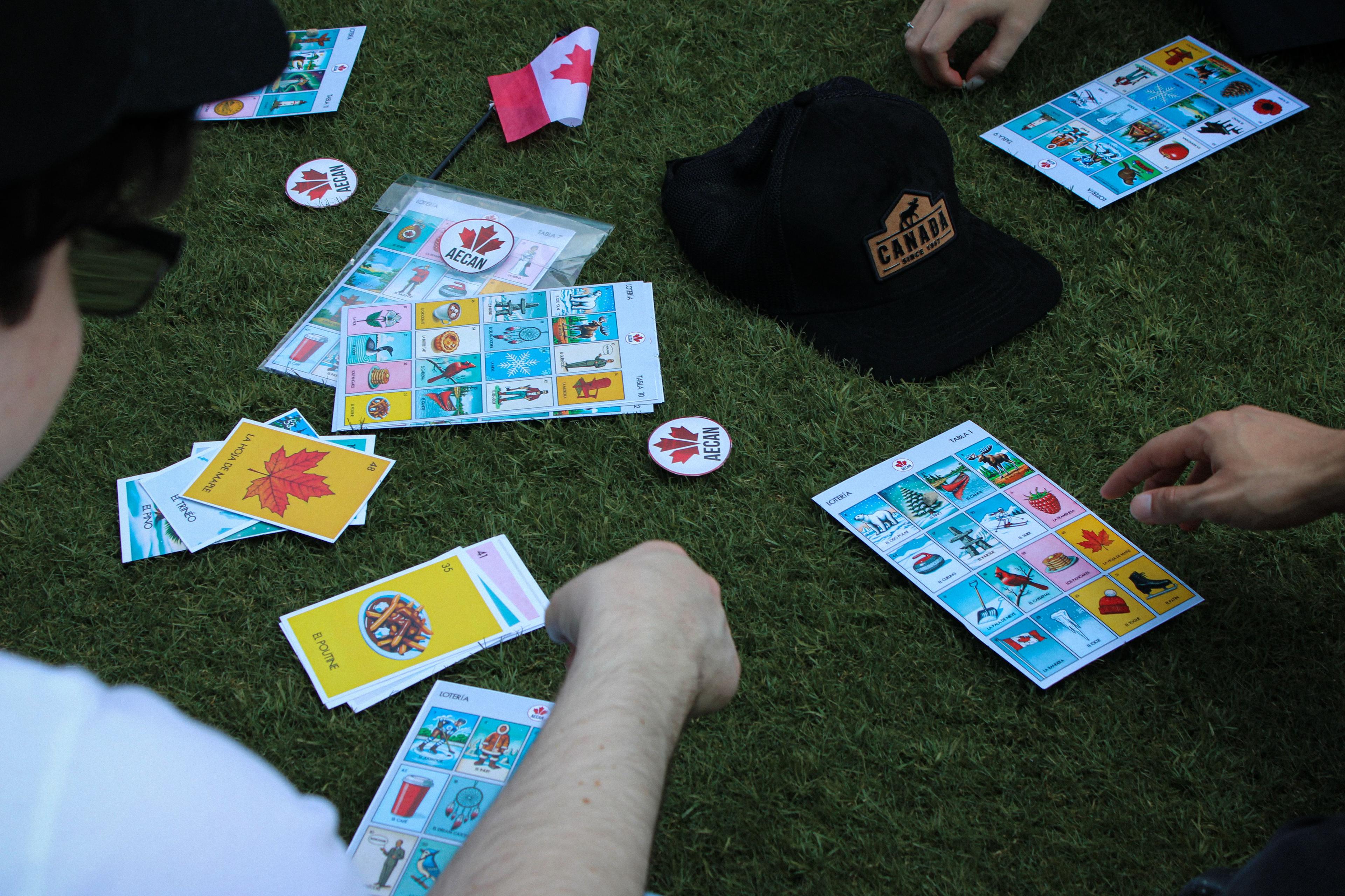 Over-the-shoulder view of a Lotería game: printed tablas, the card deck and AECAN maple-leaf chip markers on the grass